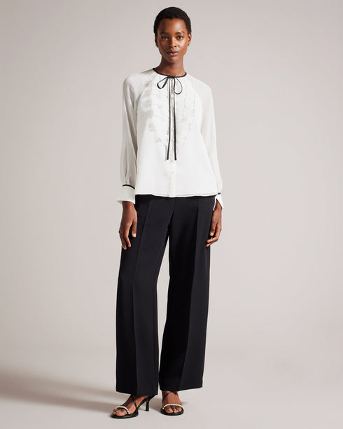 KELEIH-Tops-Channel vintage glamour with a modern twist in the KELEIH blouse. Its delicate organza ruffles and darts exude timeless elegance, while the crew neck keeps it versatile. Whether you're attending a formal event or dressing up for a date night, this blouse will effortlessly elevate your look. Blouse Organza ruffles on front Long sleeves Contrast stitching and neck tie Button fastening Darts on shoulders Crew neck with contrast binding Silvia is 5'11" / 1.8m and wears White in a Ted size 1- Ted Bak